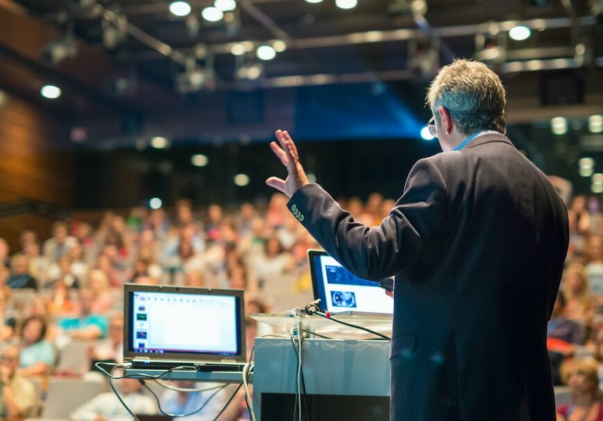 conference-speaker-stage-M A man on stage delivering a presentation.